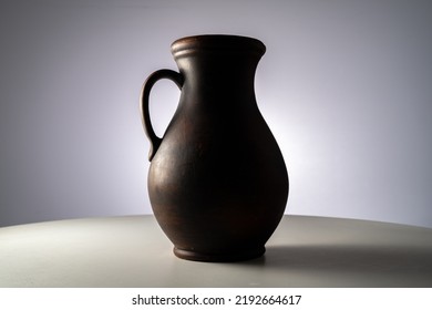 Brown Earthenware Jug With A Handle In A Dark Studio. An Old Jar From Clay Standing On A White Table Isolated On A Gray Background. Handmade Ancient Pitcher For Drinks And Food. Old Rustic Cookware.