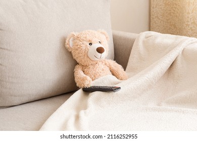 Brown fluffy teddy bear sitting on beige sofa under blanket with remote control and watching tv. Home relax. 