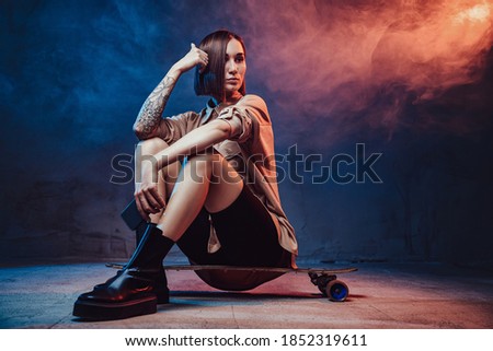 Brown haired beautiful girl with tattoo and smartphone poses in smokey background sitting on skateboard.