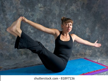 A brown haired caucasian woman is smiling doing yoga exercise, Navasana Pose or Rotated Boat posture  on yoga mat in studio with mottled background.