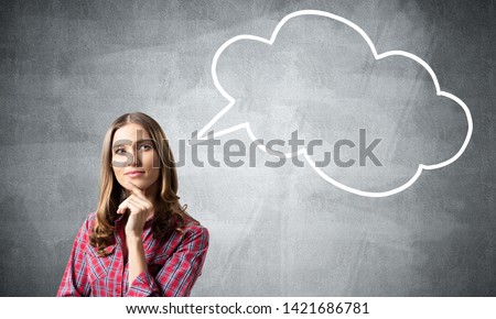 Brown haired woman looks pensive upwards and tries to remember something. Blank speech bubble illustration on grey wall. Puzzled girl has serious facial expression. Woman wears red checkered shirt