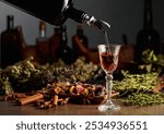 Brown herbal liquor or balm is poured from a vintage bottle into a glass. On a table dried herbs, flowers, spices, and old kitchen utensils. Concept of herbal medicine.