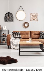 Brown Leather Couch With Blanket And Patterned Cushions In Real Photo Of Bright Living Room Interior With Coffee Table With Lemon Water And Book And Mirror, Clock And Poster On The Wall