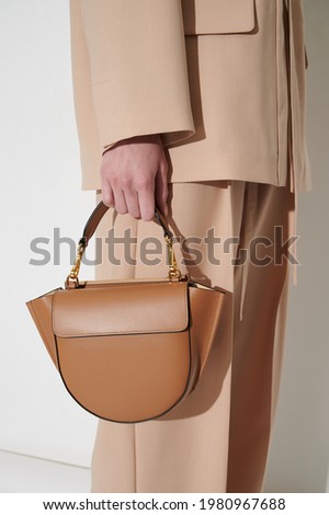 Brown leather handbag in girl's hand. Person with luxury bag in beige classic suit. Vertical photo.