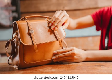 A brown leather handbag is sitting on a wooden table. A woman's hands are opening the bag.