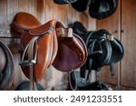 brown leather saddle hanging on wooden wall at stable or barn, professional equestrian horse riding equipment