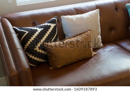 Brown leather sofa with graphic pattern pillow in elegant living room interior by the window. Modern stylish interior.