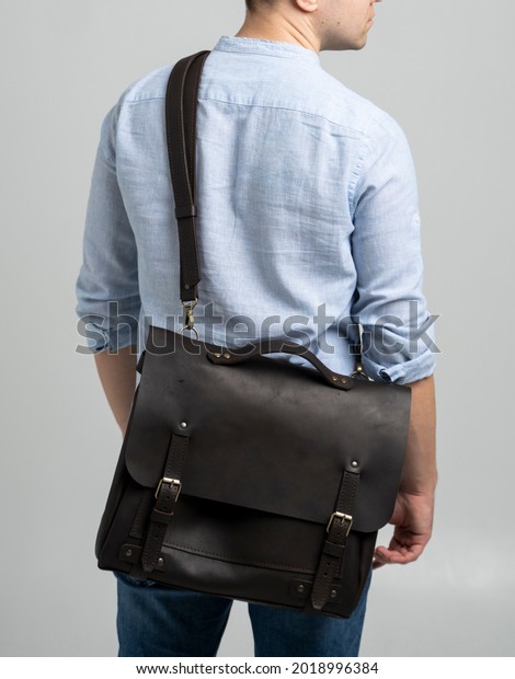 Brown men's shoulder leather bag for a
documents and laptop on the shoulders of a man in a blue shirt and
jeans with a white background. Satchel, mens leather handmade
briefcase.