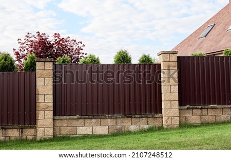 Brown metal profile fence with block posts. Incline construction. Corrugated surface. Security. Private property fencing. Opaque hedge. Outdoor house exterior. Side view. Urban or industrial style.