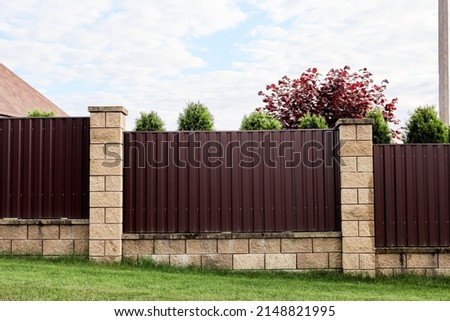 Brown metal profile fence with block posts. Incline construction. Corrugated surface. Security. Private property fencing. Opaque hedge. Outdoor house exterior. Side view. Urban or industrial style.