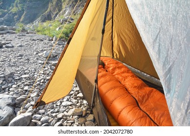 Brown One Person Hiking Pole Tent With Orange Down Sleeping Bag Inside Camping On River Rocks. Close Up Of Mesh Door Open.