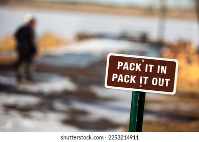 a brown, pack it in, pack it out sign with white lettering and white boarder. Taken at Cheery Creek reservoir in Colorado. With snow trees and a person walking in the background. - Shutterstock ID 2253214911