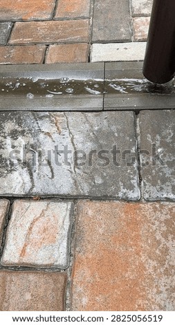 Brown rain gutter downspout during rainfall, with water flowing onto paving stones and collecting in a shallow depression near the drain. Close-up of rainwater drainage and wet pavement.
