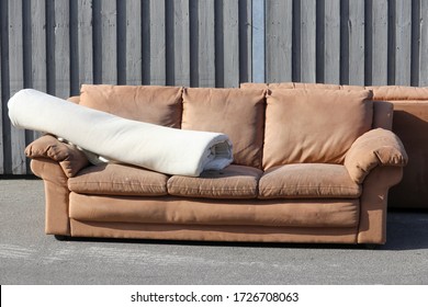 A  Brown  Second Hand Used Couch Standing Outside On Sidewalk. A Rolled Up Carpet Is Laying On The Sofa. The Furniture Is Old And Worn.