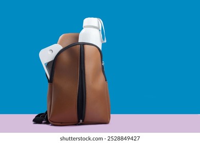 Brown shoulder bag with a water bottle and alcohol spray, ready for traveling or going to school. Bright background, blue and pink - Powered by Shutterstock