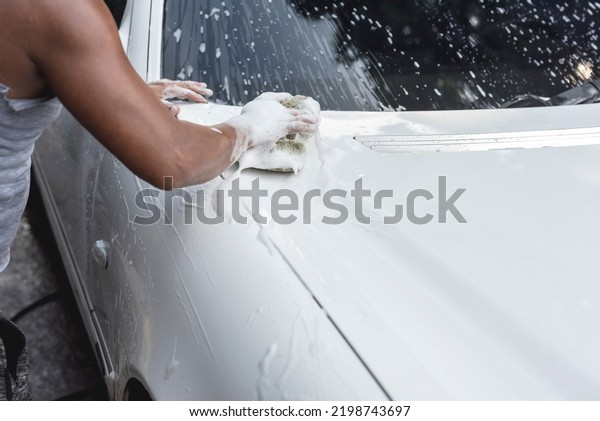 A brown skinned man in a tank top cleans\
the hood of a white sedan with a\
sponge.