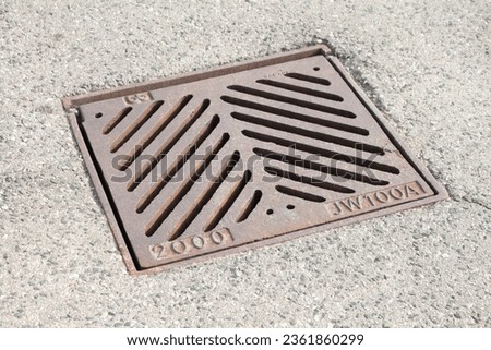 brown square sewer in between dry pavement with opposite diagonal hole lines on it that says 2000 jw100a outside exterior in summer with sun shining on it
