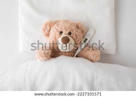 Brown teddy bear with digital thermometer lying down on pillow and sheet under blanket in white bed. Temperature measuring in cold and flu virus time. Children healthcare concept. Closeup.