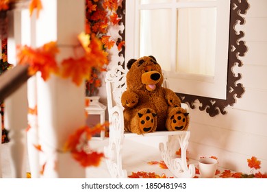 Brown Teddy bear sitting on a chair on the porch in autumn, autumn leaves around. Toys for children, warmth and comfort in the house, white porch of the house - Powered by Shutterstock