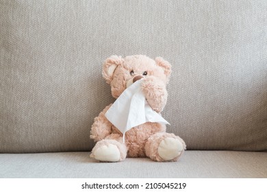 Brown teddy bear sitting on sofa and wiping nose with white paper napkin. Cold and flu virus. Children healthcare concept. Closeup. Front view.