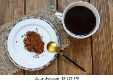 Brown Vegetable Carob (Ceratonia Silica) In Powder Form Without Caffeine. Natural Tasty Drink Made From Carob. A Cup With Hot Carob, A Saucer With Powder And A Spoon On A Wooden Background.