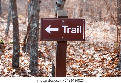 Brown and white hiking trail marker sign, in close-up, with arrow on a wooden post amid bare trees and a bed of fallen leaves in a forest. Early winter, selective focus on trail sign. - Powered by Shutterstock