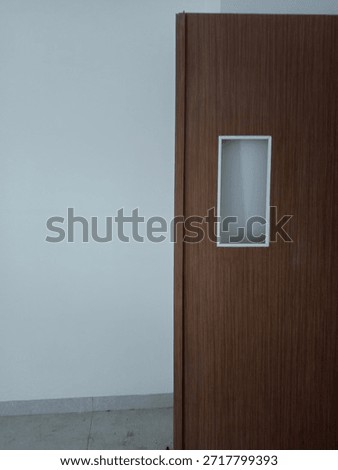 ​Modern brown wooden door with a small rectangular glass window leaning against a white wall.