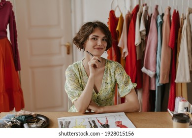 Brown-eyed curly short-haired woman in stylish floral dress smiles sincerely, holds pen and poses in cozy office of fashion designer.