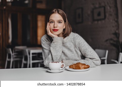 Brown-eyed Girl In Woolen Sweater Leaned On White Table In Cafe And Looking At Camera. Photo Of Woman With Red Lips Ordering Coffee And Croissant