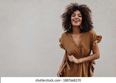Brown-eyed woman in brown dress smiles on grey background. Stylish brunette curly dark-skinned lady moves on isolated.