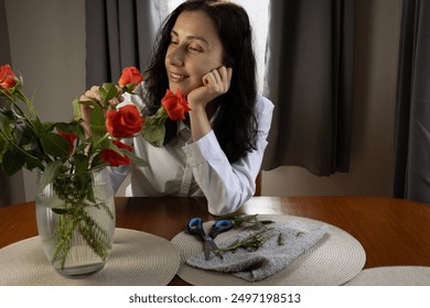 Brown-haired girl admires a bouquet of flowers. Young woman looks at red roses. Flower arrangement in a vase. - Powered by Shutterstock