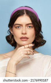 A brown-haired girl in a white blouse, rings, a bracelet and golden earrings is posing on the blue background. The girl's hair is fixed with a wide violet headband cover - Shutterstock ID 2176990187
