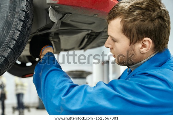 Brown-haired man with light stubble examining car\
suspension of lifted automobile . Competent expert in blue uniform\
and black gloves fixing broken parts of the car at working\
place