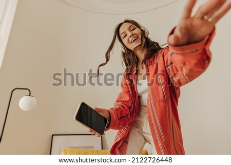 Brownhaired with wavy hair listens to music through headphones and holds phone, dressed in casual clothes. Cute caucasian model has fun at home alone in free time. Modern gadgets or phone use concept. Stock photo © 