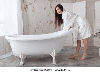 Brown-haired Young Woman In A Bathrobe, Standing In White Room And Preparing Buble Bath