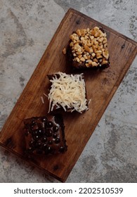 Brownie Cake Laid Out On A Wooden Cutting Board. Brownie Cake Topping With Nuts, Cheese And Choco Chips. Closeup Photo.