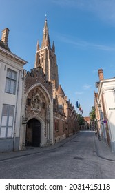 Brugge, Flanders, Belgium - August 4, 2021: Sunlit Onze Lieve Vrouw Cathedral tower under blue sky behind monumental entrance gate to Gruuthuse palace and museum. Street scene with flags.