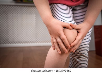Bruised Knee In A Child At Home. Boy Holding His Knee With His Hands. Knee Pain In A Child