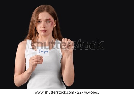 Bruised young woman holding paper with text I'M FINE on dark background. Domestic violence concept