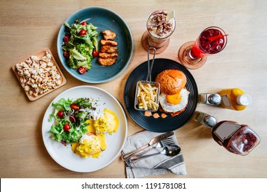 Brunch Set Including Egg Benedict, Grilled Chicken And Burger With Fried Egg With Popcorn And Beverages On Wooden Table.