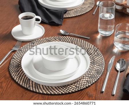 Brunch Table Setting Featuring Elegant White Porcelain Dishware on Woven Rattan Chargers, Stainless Steel Flatware and Clear Glass Drinkware, Freshly Brewed Cup of Coffee on a Rich Wooden Dining Table