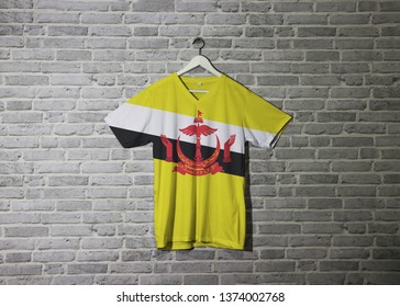 Brunei Darussalam flag on shirt and hanging on the wall with brick pattern wallpaper, red crest on yellow field cut by black and white diagonal stripes.