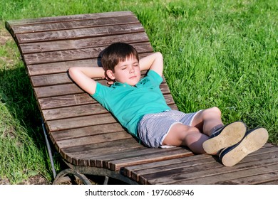 Brunet Boy Of 8 Years In T-shirt And Shorts Lies On Wooden Deck Chair In The Sun Against The Backdrop Of A Green Lawn. The Boy In Relaxed, Lazy Position, Closed His Eyes.