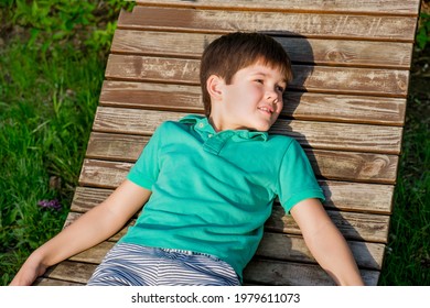 Brunet Boy Of 8 Years In T-shirt Lies On Wooden Deck Chair In The Sun Against The Backdrop Of A Green Lawn. The Boy In Relaxed, Lazy Position, Closed His Eyes.