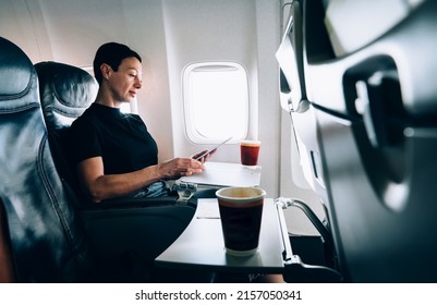 Brunette 40 Years Old Female Airplane Passenger With Tablet In Hands Sitting In Comfortable Seat Near The Window. Attractive Woman Enjoying Flight While Using With Touch Pad Online Connection On Board