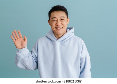 Brunette Adult Asian Man Smiling And Waving At Camera Standing Isolated Over Blue Background