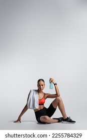 brunette african american woman in sports bra and bike shorts sitting with sports bottle and towel on grey background