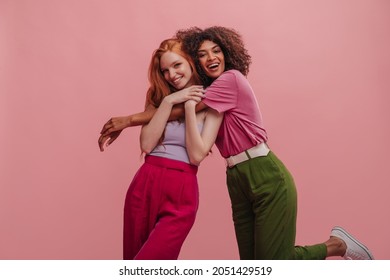 Brunette with afro hair hugs embarrassed girlfriend with red hair on pink background. Beauties in casual bright clothes express positive emotions. Beauty, people emotions and summer vacation concept. - Powered by Shutterstock