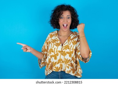 Brunette Arab Woman Wearing Hawaiian Shirt Over Blue Background Points At Empty Space Holding Fist Up, Winner Gesture.