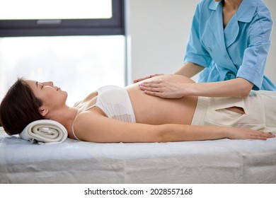 Brunette asian pregnant woman receiving osteopathic treatment in spa cabinet in clinic. Manual therapist manipulates woman's belly, carefully professionally doing various techniques. side view - Powered by Shutterstock